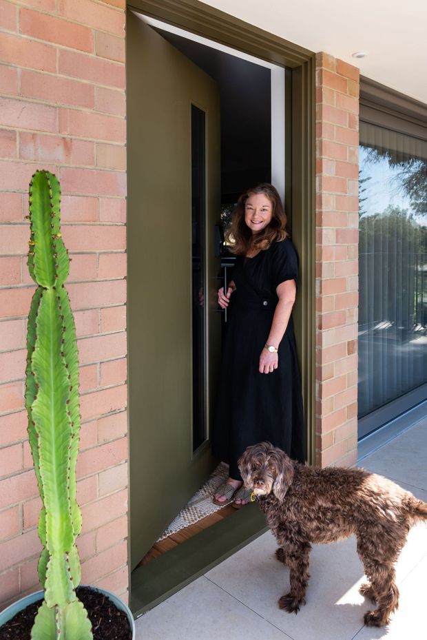 A residential entryway viewed from outside, featuring a dark green front door set within a pinkish-red brick facade. A woman stands in the open doorway with a dog, while a tall potted cactus sits beside the entrance. A large adjacent window with vertical blinds reflects outdoor trees and bright daylight.