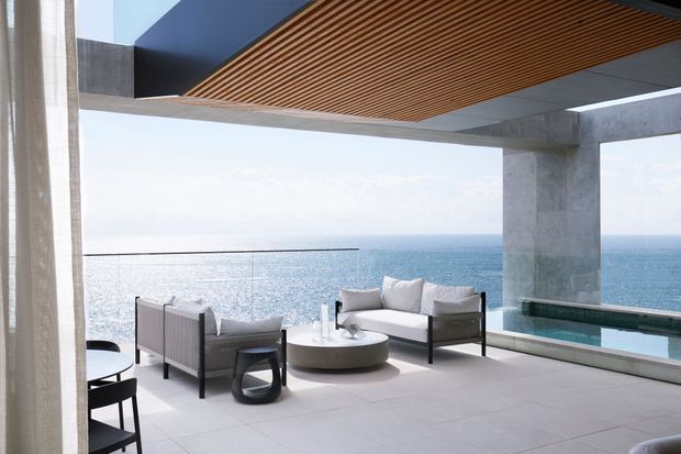 A refined contemporary oceanfront terrace or outdoor lounge framed by exposed concrete columns and expansive glazing. The space features a pale tiled floor, minimalist outdoor seating, a round coffee table, a timber slatted ceiling, and a narrow integrated pool or water feature along the seaward edge. The open view directly overlooks the ocean.