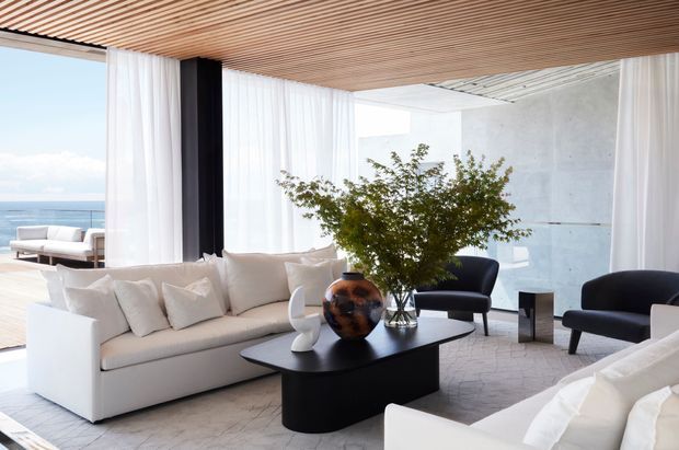 Bright contemporary living room with a large cream sectional sofa, dark sculptural coffee table, black lounge chairs, white curtains, and a leafy indoor tree. A slatted timber ceiling adds warmth, while floor-to-ceiling glazing opens toward a coastal view and floods the room with natural light.