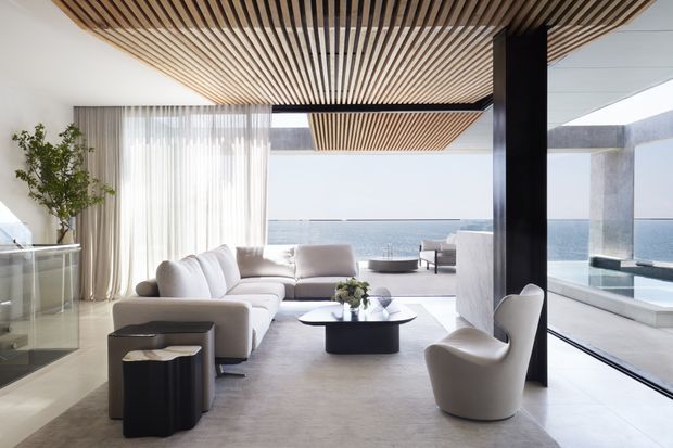 A bright, sophisticated contemporary living room opening onto a waterfront terrace and pool. The space features a large cream sectional sofa, sculptural lounge chair, low dark coffee table, sheer floor-to-ceiling curtains, expansive glazing, pale flooring, and a striking slatted timber ceiling with a dark structural column.