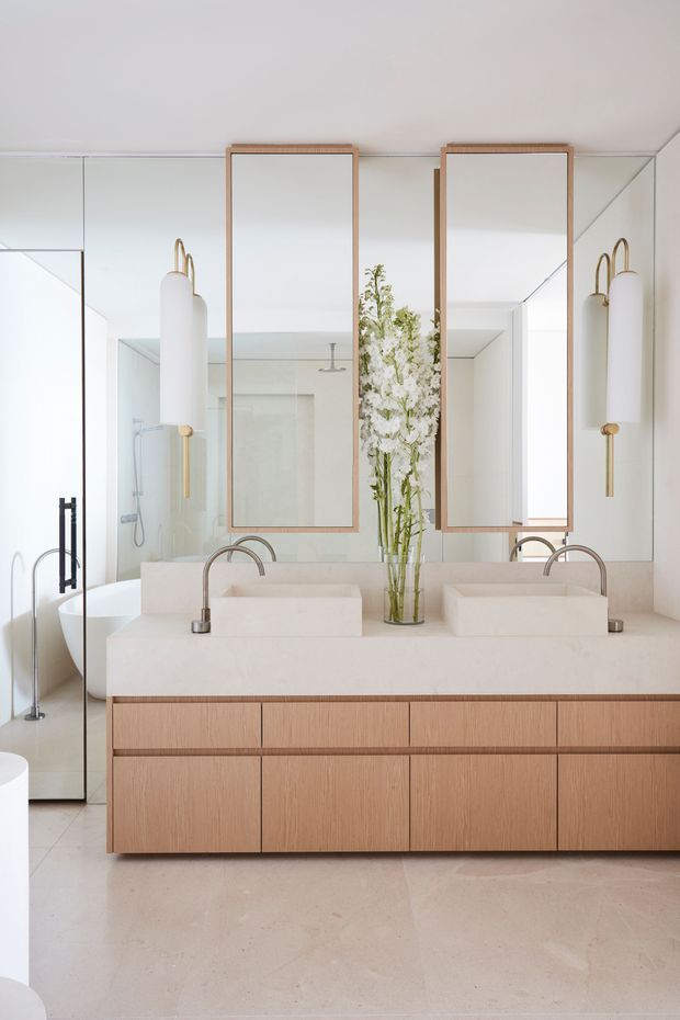 Bright contemporary bathroom featuring a double floating timber vanity with a pale stone benchtop, twin integrated basins, curved brushed-metal tapware, large timber-framed mirrors, and sculptural wall sconces. A glass-enclosed shower and freestanding fixtures are visible in the mirrored and side views. The room has a refined, warm-neutral palette and reflective surfaces.
