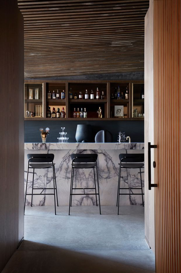 A sophisticated home bar viewed through a partially open timber door. The room features a long veined marble bar front and benchtop, dark built-in timber shelving filled with bottles and glassware, three black bar stools, a dark backsplash, and a slatted timber ceiling and door. The atmosphere is moody, refined, and contemporary.