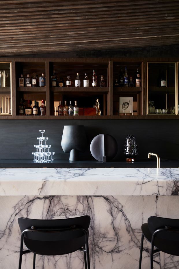 A sophisticated home bar with dark timber open shelving filled with bottles and glassware, a black backsplash and counter, a dramatic veined marble island with waterfall sides, and black bar stools. Sculptural decorative objects, a crystal glass display, and a brushed gold tap add refined contrast. The ceiling features dark timber slat detailing.
