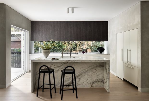 Contemporary kitchen with a large marble island and two black bar stools, dark timber overhead cabinetry, pale built-in cabinetry, and a wide window above the benchtop. The room has light timber flooring, smooth plastered walls, minimalist track lighting, and a glazed door opening to an exterior balcony or deck.