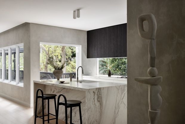 Contemporary kitchen with a large marble-look island and waterfall end, black bar stools, integrated dark cabinetry, concrete-toned walls, white-framed windows, and a sculptural wall-mounted feature. The space is bright and connected to leafy outdoor views.