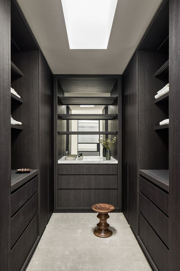 A narrow, highly organised walk-in wardrobe lined with dark timber-look built-in cabinetry, open shelving and drawers on both sides. A central vanity with a light stone benchtop, drawers and mirror is positioned at the end, with a skylight above and a small sculptural wooden stool in the aisle. The space has a dark, refined, contemporary feel with strong symmetry and integrated lighting.