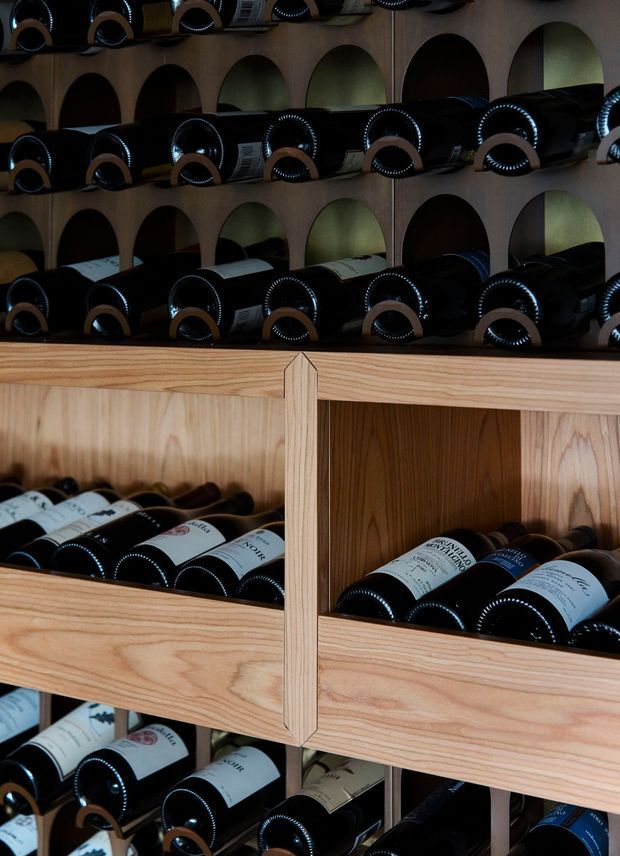 Close-up view of a built-in wine storage display filled with horizontally stacked wine bottles. The upper section features dark arched cubby openings with concealed or low lighting, while the lower section is constructed from warm natural timber with open bottle compartments and vertical dividers. The image is tightly framed around the joinery and bottles, creating a dark, refined cellar-like atmosphere.