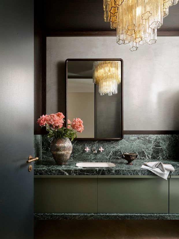 A sophisticated bathroom vanity viewed through a partially open dark door. The room features a long floating olive-green vanity with a richly veined dark green marble benchtop and integrated backsplash, a white basin, wall-mounted pink-toned tapware, and a rounded-rectangle framed mirror. A sculptural tiered glass-and-gold pendant light hangs above and is reflected in the mirror. Decorative flowers and a folded towel add softness, while dark timber framing and warm low lighting create a luxurious, moody atmosphere.