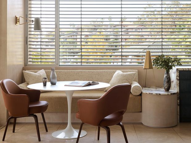 A refined dining and seating nook with a built-in upholstered banquette beneath large horizontal louvred windows. A white pedestal table is paired with two brown leather dining chairs, with neutral textured upholstery, a sculptural floor lamp, ceramic accessories, and a large cylindrical side table creating a warm mid-century-inspired interior.