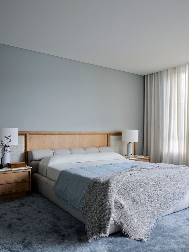 A calm, contemporary bedroom with a large upholstered bed set against a built-in light timber headboard. Soft grey-blue walls, floor-to-ceiling sheer curtains, bedside tables with lamps, and layered blue-grey bedding create a muted, refined atmosphere. A dark textured carpet covers the floor.