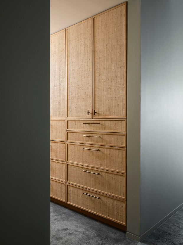 A tall built-in storage wall with cane or rattan-fronted cupboard doors and drawers, framed in warm natural wood. The joinery is set against smooth pale grey walls and a dark grey carpeted floor, with slim metal handles and a narrow passage-like setting.