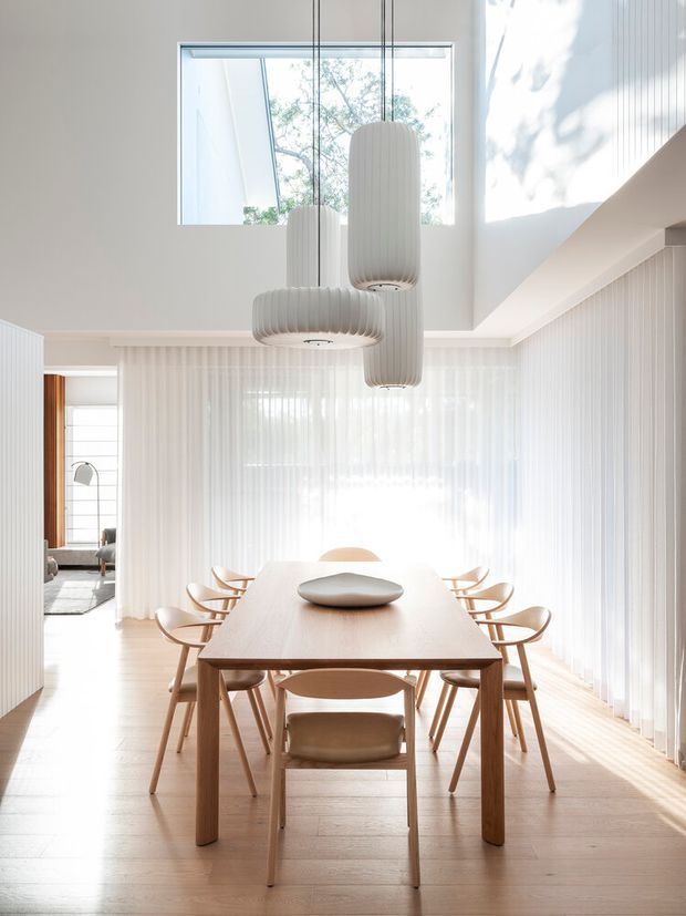 Bright, refined dining room with a long light-wood dining table surrounded by sculptural wooden chairs. Floor-to-ceiling sheer curtains diffuse abundant daylight, while three ribbed white pendant lights hang beneath a high ceiling. Vertical wall panelling, pale timber flooring, and a glimpse of an adjacent sitting area create a calm, minimalist interior.