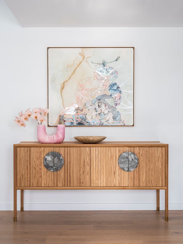 A refined interior vignette featuring a mid-century-inspired timber sideboard with vertical wood grain, slim legs, and circular stone-like door pulls. A pink ceramic vase with flowers and a shallow wooden bowl sit on top, beneath a framed abstract artwork. The palette is soft and warm against a white wall and timber floor.