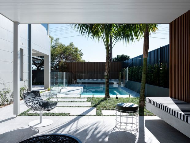 A contemporary outdoor courtyard and pool area viewed from beneath a covered patio. The space features pale tiled paving, stepping slabs through planted gravel, a compact swimming pool with glass balustrade, palm trees, dark vertical screening, and built-in striped bench seating. Modern outdoor furniture and strong sunlight create a crisp resort-like setting.