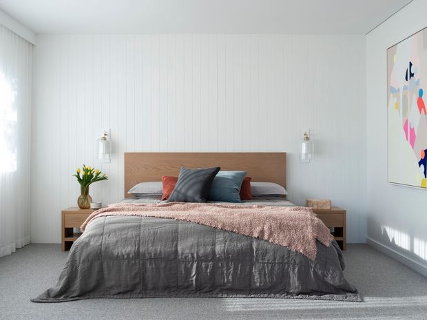 A bright, calm contemporary bedroom with a low platform bed, warm timber headboard and bedside tables, layered grey and dusty pink bedding, and white vertical wall panelling. Sheer curtains filter daylight at the window, while simple wall lights and colourful abstract artwork add restrained accents.