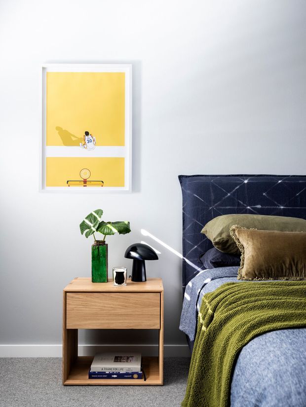 A styled bedroom vignette with a blue upholstered bed and layered olive and taupe bedding beside a light timber bedside table. The table holds a black dome lamp, green glass vase with leafy plant, and a small cup, while a framed yellow artwork hangs on the pale grey wall. Grey carpet and white skirting complete the scene.