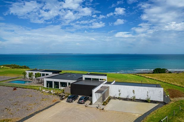 Aerial exterior view of a low-profile contemporary coastal residence set on a broad grassy site beside the ocean. The home comprises several connected flat-roofed volumes with dark and light exterior finishes, extensive glazing, paved driveways, a small swimming pool, and attached carport/garage areas. The image is dominated by the building exterior, ocean horizon, and open coastal landscape under a bright partly cloudy sky.