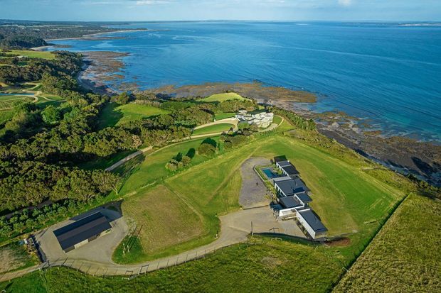 Aerial view of a large coastal property on a green, landscaped headland. Several modern low-rise buildings with dark roofs sit among expansive lawns, trees, curved driveways and gravel areas, with rocky shoreline and open blue water beyond.