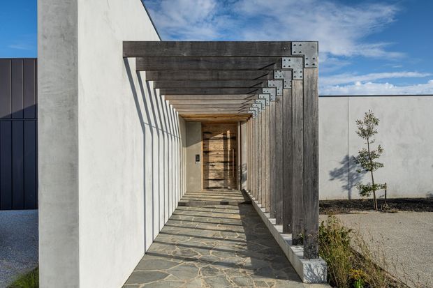 A contemporary architectural entry walkway framed by a repeated timber pergola structure. The passage has white rendered walls, dark vertical glazing at left, weathered timber beams and posts with exposed metal brackets, and an irregular natural stone-paved floor. Strong linear shadows create a dramatic, rhythmic perspective toward a timber-clad doorway, with a small landscaped courtyard visible to the right.