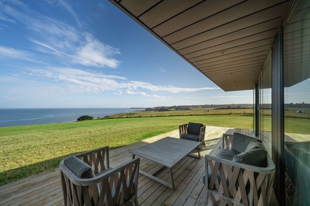 A covered timber deck overlooking a broad grassy coastal landscape and the ocean. The outdoor seating area includes woven lounge chairs and a rectangular outdoor table, while large reflective glazing and a deep soffit frame the right side of the image. The scene has a clean, contemporary coastal character with expansive views and abundant natural light.