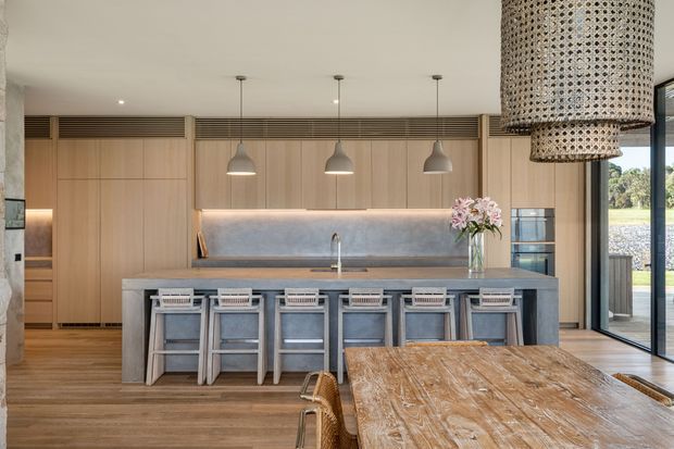 A spacious contemporary kitchen and dining area with a long timber-clad wall of integrated cabinetry, a large stone island with multiple stools, a softly illuminated backsplash, pendant lights, and a rustic timber dining table with woven chairs. Full-height glazing opens toward a landscaped exterior, while a large woven pendant adds texture overhead.