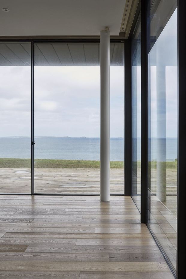 A minimalist interior opening onto a broad ocean view. The room has full-height glazed sliding doors with dark slim frames, a pale timber floor, a white cylindrical structural column, and a recessed ceiling detail. A timber deck and narrow strip of coastal landscape sit beyond the glazing, with the sea and horizon visible in the distance.