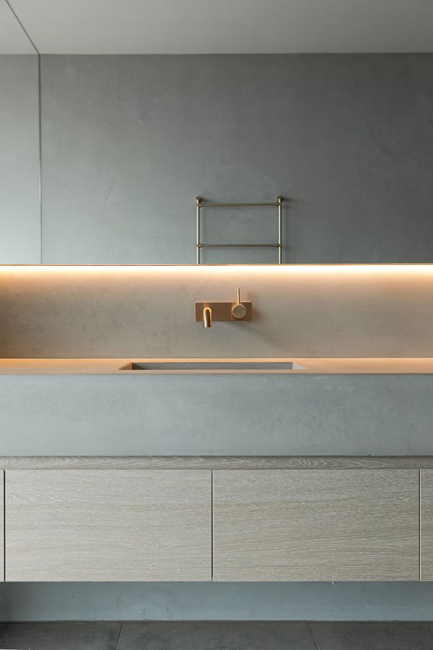 A minimalist contemporary bathroom vanity viewed straight on. The room features a broad grey microcement or concrete-look wall and benchtop, an integrated rectangular basin, brushed brass wall-mounted tapware, a slim brass towel rail, warm concealed LED lighting, and light wood-grain vanity cabinetry. Dark large-format floor tiles are visible at the bottom.