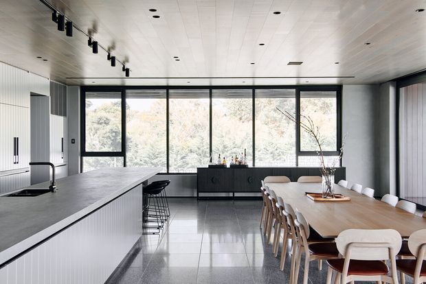 A bright, contemporary open-plan kitchen and dining area with a long island, integrated cabinetry, full-height black-framed windows, and a large timber dining table. The room features pale vertical wall panelling, a white panelled ceiling, polished light-grey tiled flooring, and minimal black track lighting. A forested view is visible beyond the glazing.