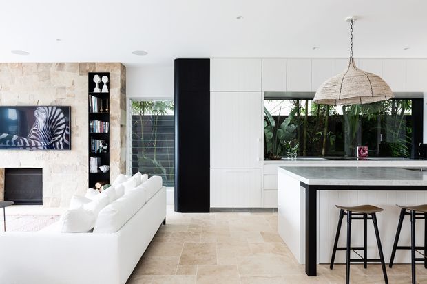 Bright open-plan contemporary kitchen and living area with white built-in cabinetry, a large stone island with bar seating, pale stone flooring, a textured stone feature wall, and abundant greenery visible through the windows. A woven pendant light adds a natural accent above the island.