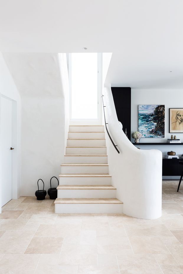 Bright contemporary interior featuring a sculptural curved staircase with white plaster balustrade, pale timber-toned stair treads, and a large vertical window at the landing. The staircase rises from a spacious light stone-tiled entry area, with a partially visible dark built-in display and artwork in the adjacent room.