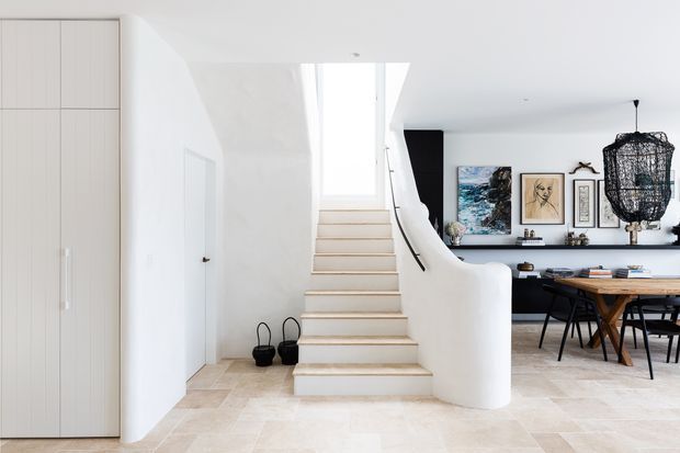 Bright contemporary open-plan interior featuring a sculptural curved staircase with pale timber treads, white walls and cabinetry, pale stone or tile flooring, and a dining area visible to the right. A black feature wall, artwork, and an oversized woven pendant add contrast and texture.