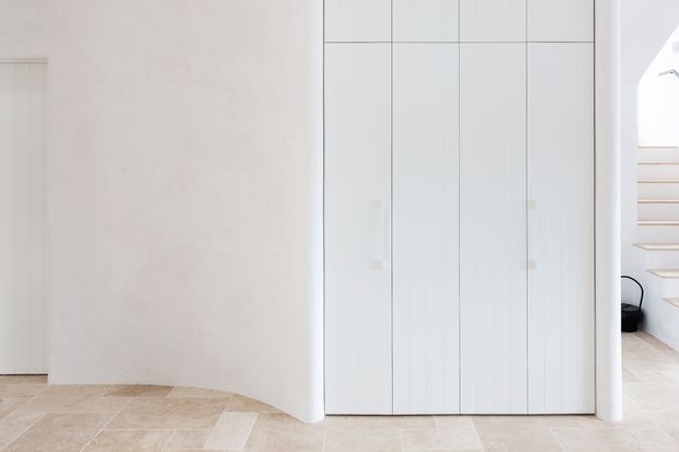 Bright minimalist interior featuring a full-height white built-in cabinet with slender vertical panel detailing, curved plastered wall returns, pale stone or limestone-look flooring, and a glimpse of an open floating staircase at right.