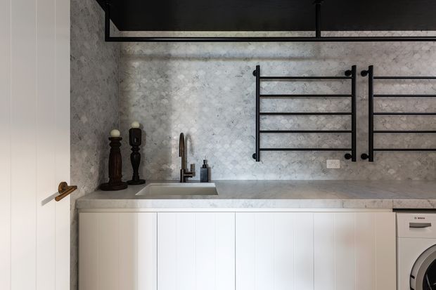 A compact contemporary laundry with white vertical-panel cabinetry, a pale stone benchtop and integrated sink, textured light stone wall finish, black wall-mounted drying racks, and a front-loading washing machine. Black accents and a dark overhead feature add contrast.