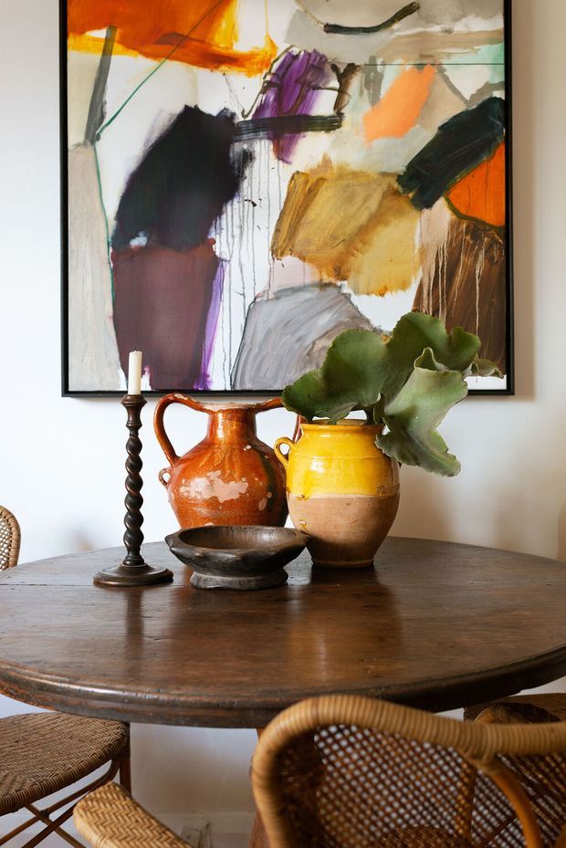 A warm dining area featuring a round dark timber dining table with woven rattan chairs, a vintage candlestick, ceramic vessels, and leafy greenery. An expressive abstract artwork in a black frame forms the backdrop against a light wall.