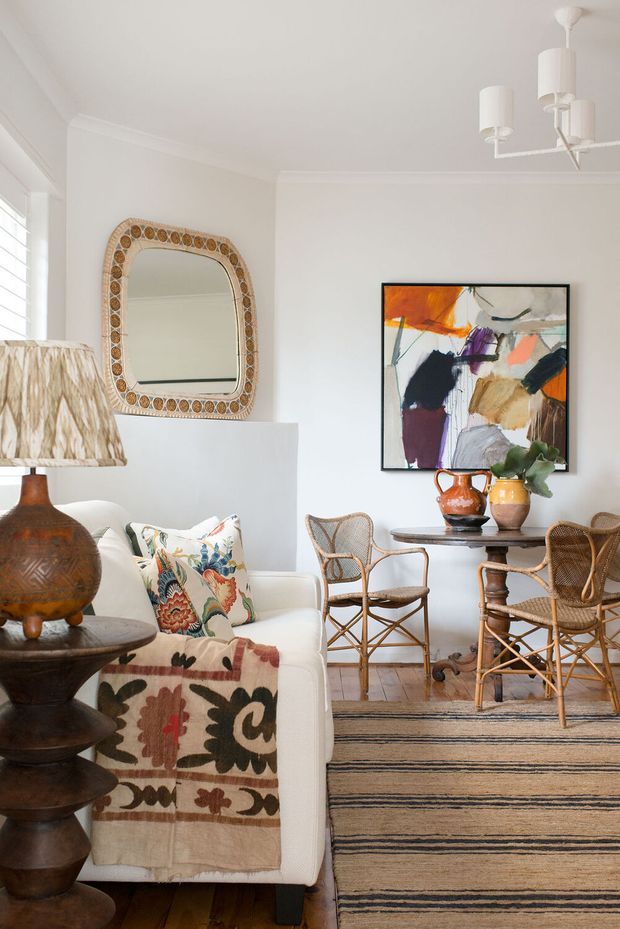 A bright, refined living and dining area with white walls, decorative ceiling moulding, shuttered windows, a white sofa, woven rattan dining chairs, a dark timber dining table, a patterned rug and throw, an ornate woven-framed mirror, and colourful abstract artwork. The room combines natural textures, vintage-inspired furniture, and eclectic decorative accents.