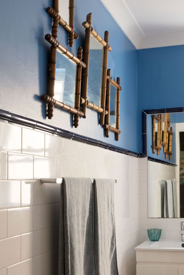 Bright bathroom interior with vivid blue painted upper walls, glossy white subway-tile wainscoting, dark trim, and bamboo-framed mirrors. A white vanity with basin and grey towels completes the space, creating a playful, eclectic look with strong natural light.