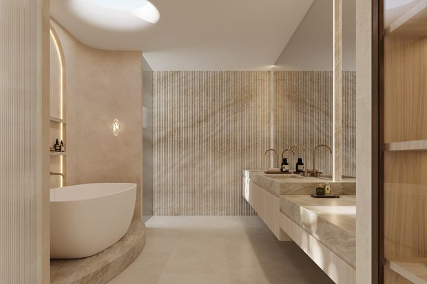 A luxurious contemporary bathroom viewed from the doorway, featuring a freestanding oval bathtub on a curved stone platform, double vanity with pale stone benchtops and integrated basins, warm timber cabinetry, brushed metal tapware, and a full-height vertically ribbed feature wall. Soft concealed lighting and a sculptural ceiling detail create a warm, refined atmosphere.