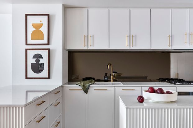 Bright, refined kitchen with white Shaker-style cabinetry, long brass pulls, a dark contrasting splashback, pale stone benchtops, and a ribbed island front. Framed geometric artwork and restrained styling add a contemporary classic character.