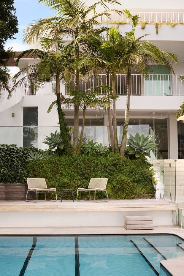 A bright multi-storey white residential building viewed from a landscaped pool area. The facade features broad glazing, white vertical railings, balconies, and a flat roofline with trailing greenery. Tropical palms, sculptural agaves, dense hedging, a timber-toned pool deck, and a turquoise swimming pool create a resort-like outdoor setting.