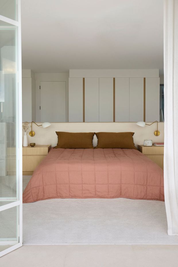 A bright, calm bedroom viewed through open glazed doors. The room features a low upholstered bed with a dusty rose quilt and brown pillows, light timber bedside cabinetry, brass wall-mounted reading lamps, pale carpet, and a full-width built-in wardrobe with white slab doors and narrow timber accents. The palette is soft and warm with clean contemporary detailing.