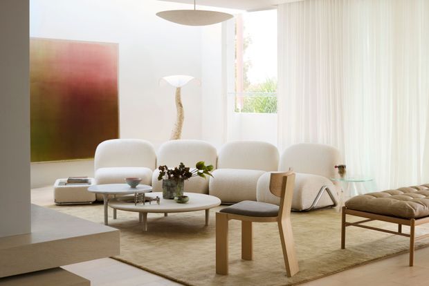 Bright, softly styled contemporary living room with a curved cream sofa, sculptural lounge chairs, layered round coffee tables, a timber armchair, and a tufted bench. Floor-to-ceiling sheer curtains and large windows provide abundant natural light, while a large abstract artwork and sculptural lighting add refined focal points. The room has pale timber flooring and a neutral, warm-toned palette.