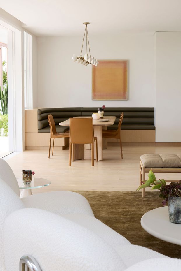 Bright, refined open-plan living and dining area with a built-in black banquette, round timber dining table, warm tan dining chairs, pale timber flooring and full-height glazing. A sculptural pendant light and abstract artwork add understated contemporary character, while a white curved sofa and low coffee table occupy the foreground.