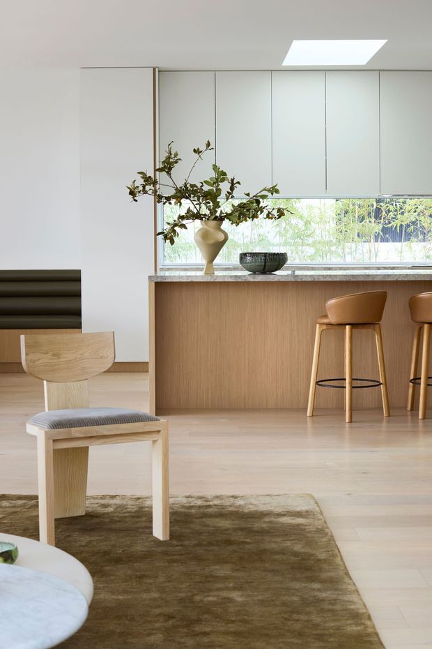 A bright, refined open-plan interior showing a kitchen with a timber-fronted island and integrated seating, opening toward a living area. Pale timber flooring, a sculptural timber chair, upholstered bar stools, white full-height cabinetry, a stone benchtop, and a large skylight create a warm minimalist setting. Indoor greenery and a garden view through the horizontal window add an organic element.