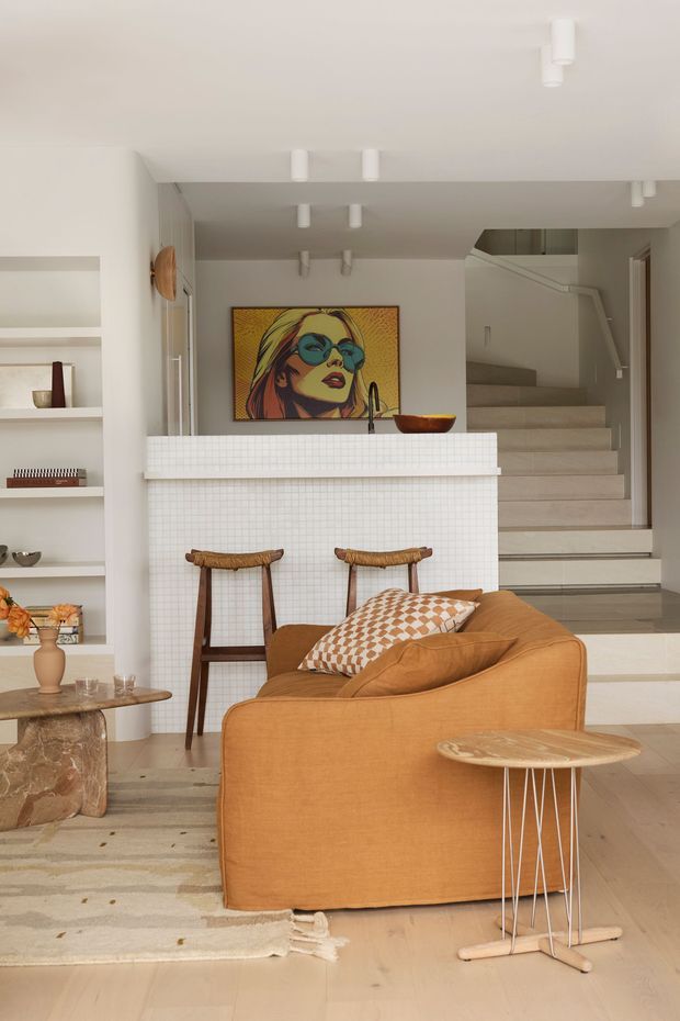 Warm contemporary open-plan living area with a rust-orange sofa in the foreground, a small round timber side table, and a chunky natural timber coffee table. Behind the sofa is a tiled kitchen island with two woven timber bar stools, open shelving, and a colourful framed pop-art portrait. Pale timber flooring, light neutral walls, and a staircase with a glass balustrade create a bright, refined interior. Recessed and surface-mounted white ceiling lights complete the space.