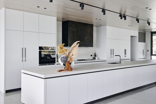 Bright contemporary kitchen with a long white fluted island, extensive full-height white cabinetry, integrated appliances, a dark stone feature panel, pale timber-look ceiling, black hardware and track lighting. The space has a clean, refined, minimalist appearance with subtle black accents.