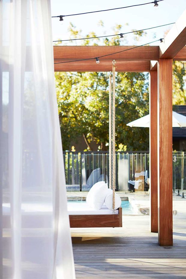 Bright outdoor entertaining area viewed through a sheer curtain, featuring a timber pergola with exposed beams, festoon string lighting, a suspended timber swing seat, timber decking, pool fencing, and a partially visible swimming pool. The space is leafy and relaxed with strong indoor-outdoor connection.