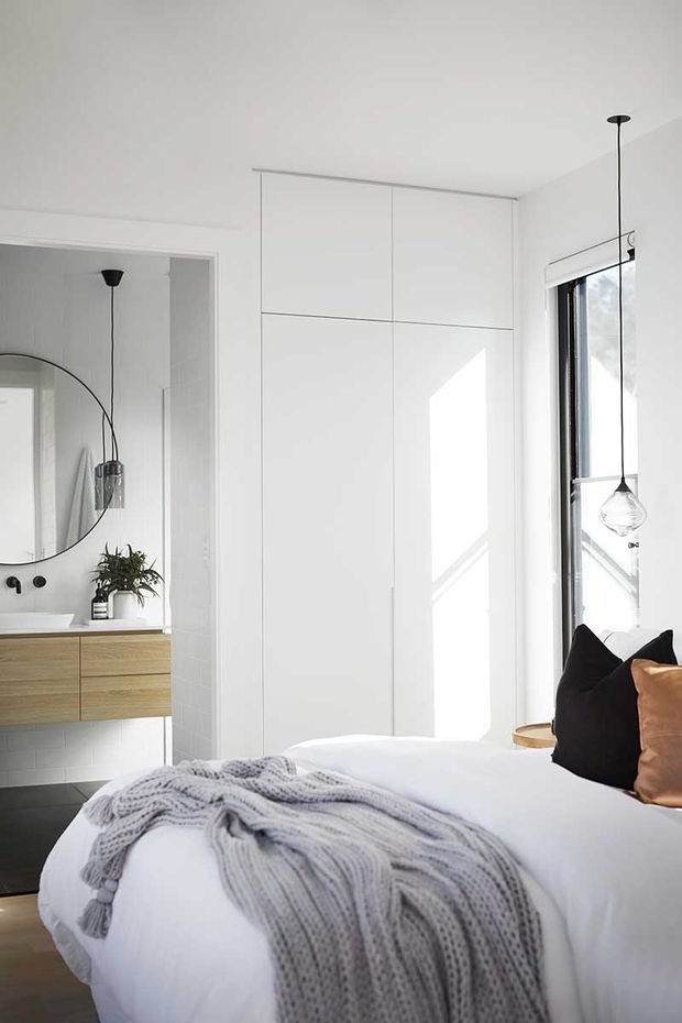 Bright contemporary bedroom with a predominantly white palette, floor-to-ceiling built-in wardrobe cabinetry, large window, and a partially visible adjacent bathroom vanity. The bed is dressed in white linen with a chunky grey knitted throw and dark and tan cushions. Minimal black pendant lights and a round wall mirror add contrast.