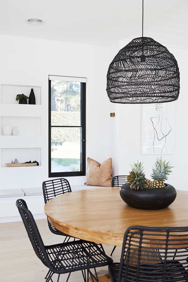 Bright contemporary dining area with a round light timber dining table, black woven dining chairs, and a large sculptural black woven pendant light. White walls feature recessed display niches, a narrow black-framed window, minimalist artwork, and a built-in white bench. A black bowl centerpiece holds pineapples and spiky foliage.