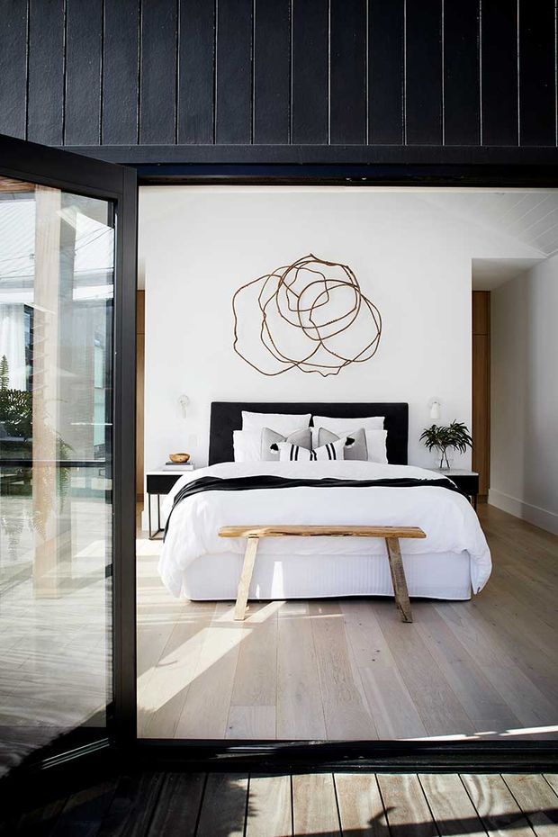 A bright, contemporary bedroom viewed through an open black-framed glass door. The room features a white upholstered bed with black headboard, layered white bedding and patterned cushions, pale timber flooring, a simple timber bench, white walls, abstract wire wall art, bedside tables, and small plants. Dark vertical timber cladding and a black ceiling/door frame create strong contrast with the light interior.