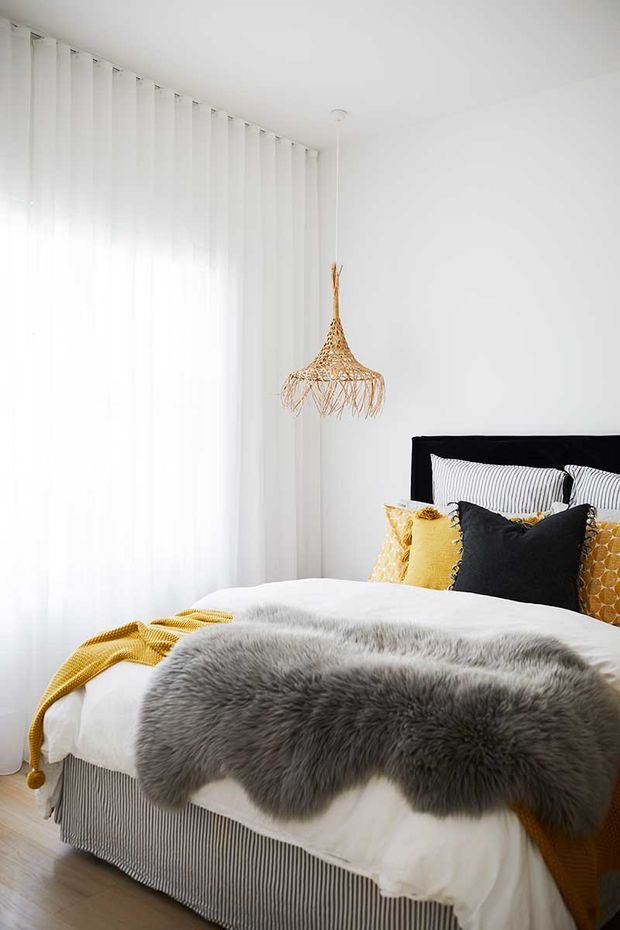 Bright, softly styled bedroom featuring a black bed frame, white bedding, a grey faux-fur throw, mustard and black accent cushions, full-height sheer white curtains, and a woven pendant light. The room has a clean, cozy contemporary feel with light timber flooring.